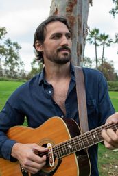 Bearded musician in a navy shirt strumming an acoustic guitar during an outdoor park session, leather strap over his shoulder with grassy lawn, palm trees and eucalyptus in the background.