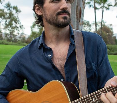 Bearded musician in a navy shirt strumming an acoustic guitar during an outdoor park session, leather strap over his shoulder with grassy lawn, palm trees and eucalyptus in the background.