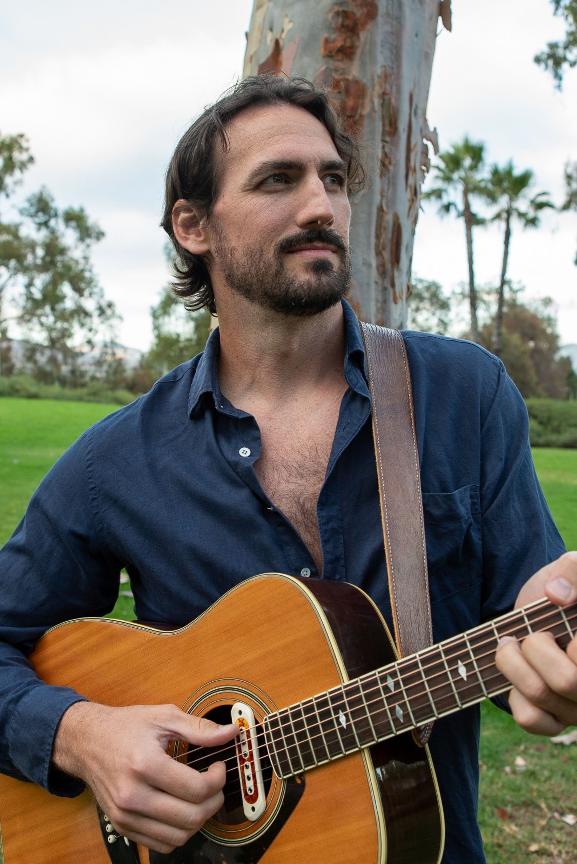 Bearded musician in a navy shirt strumming an acoustic guitar during an outdoor park session, leather strap over his shoulder with grassy lawn, palm trees and eucalyptus in the background.