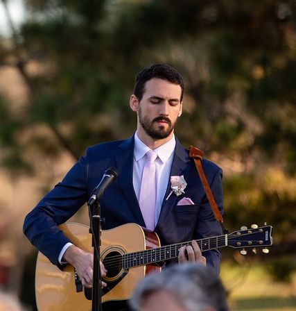 Man in navy suit with pink tie and boutonniere playing acoustic guitar into a microphone at an outdoor wedding ceremony.