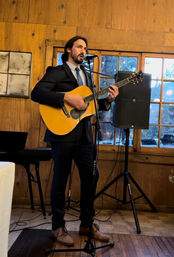 Musician in a suit playing an acoustic guitar and singing into a microphone in a rustic wood‑paneled room with windows and a speaker.