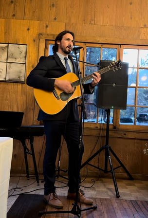 Musician in a suit playing an acoustic guitar and singing into a microphone in a rustic wood‑paneled room with windows and a speaker.