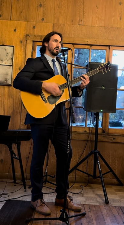 Musician in a suit playing an acoustic guitar and singing into a microphone in a rustic wood‑paneled room with windows and a speaker.