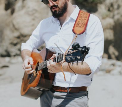 Bearded man in sunglasses strumming an acoustic guitar on a sunny coastal beach with rocky cliffs in the background