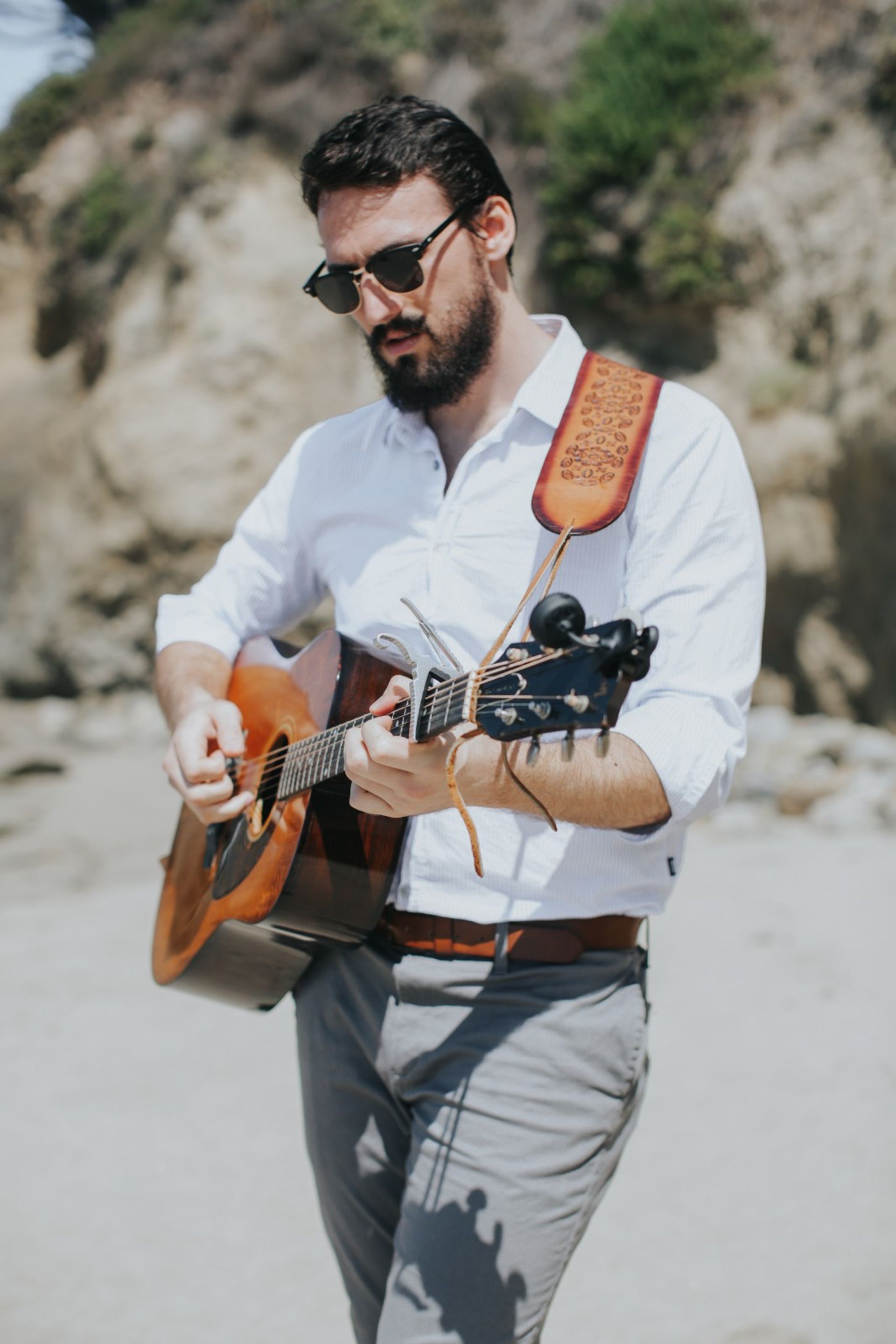 Bearded man in sunglasses strumming an acoustic guitar on a sunny coastal beach with rocky cliffs in the background