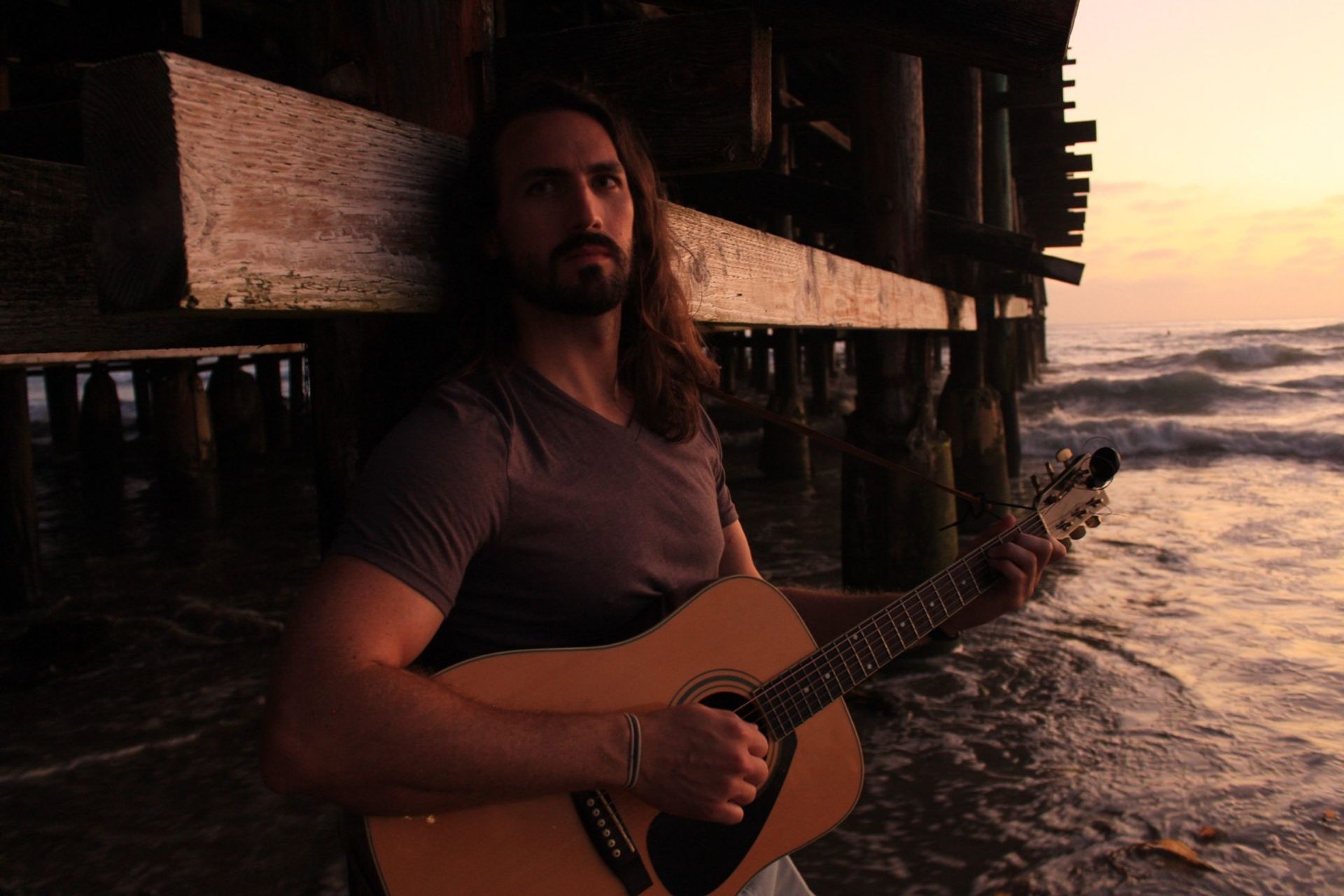 Long-haired person strumming an acoustic guitar beneath a weathered wooden pier at sunset, ocean waves lapping the shore