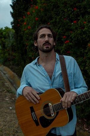Bearded man in a light denim shirt holding an acoustic guitar outdoors beside a green hedge dotted with red flowers, looking thoughtful.