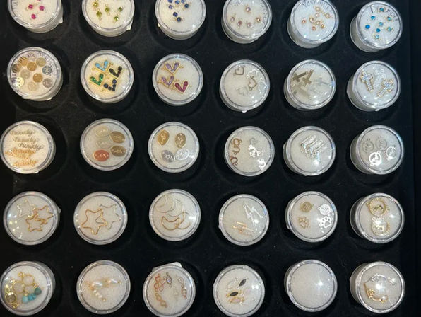 Close-up of a black display tray with an organized grid of clear round containers holding white foam and assorted tiny jewelry charms and earring studs—gold, silver and colorful rhinestone accents.