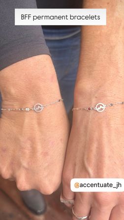 Close-up of two wrists wearing matching delicate chain BFF permanent bracelets with round mountain-cutout charms and tiny gemstone accents on silver chains, minimalist friendship jewelry in a casual outdoor setting.