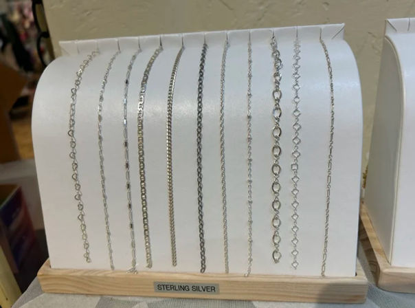 Gleaming sterling silver necklace chains in assorted link styles hung on a white curved display stand with a wooden base — retail jewelry display.