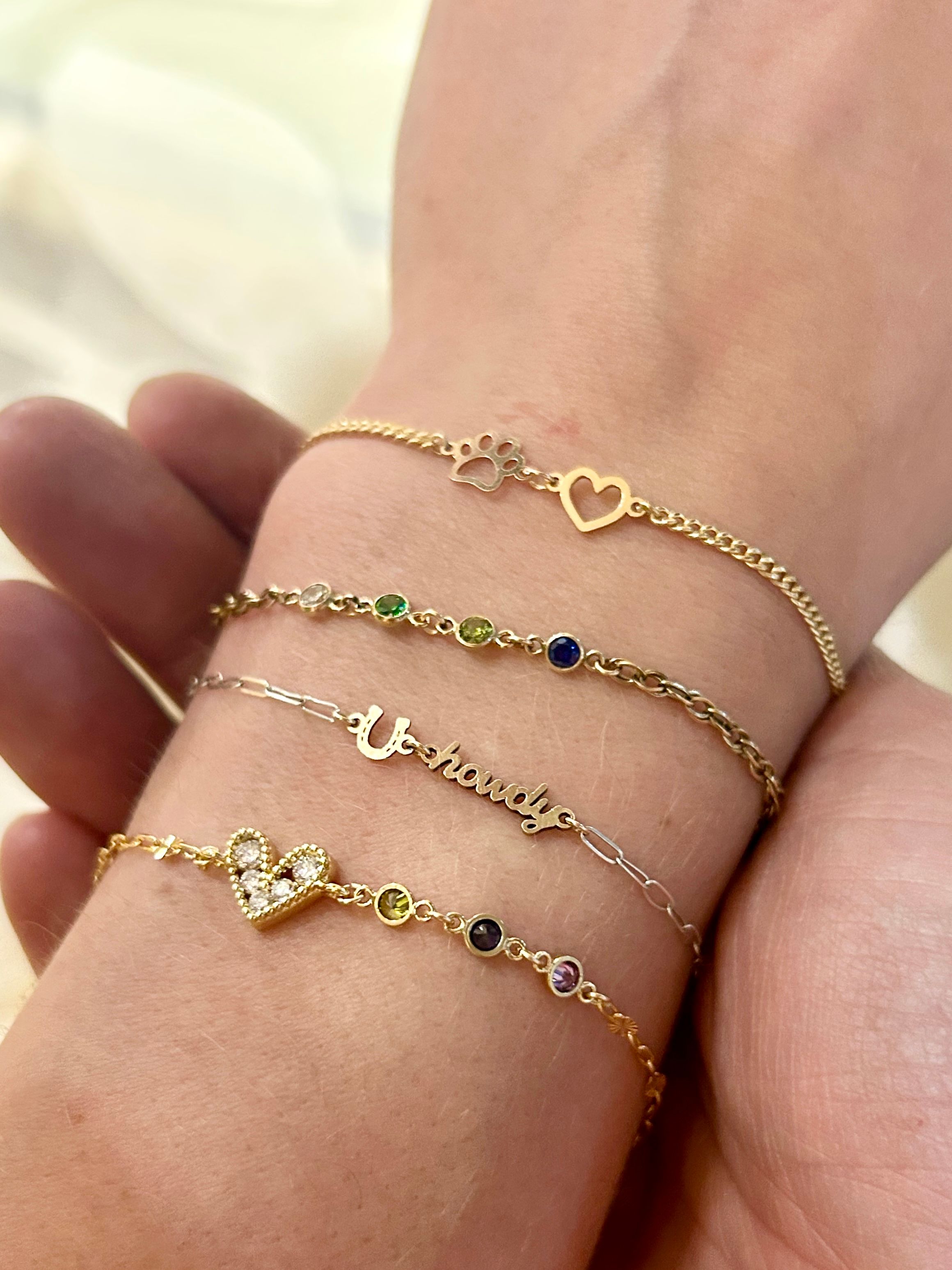 Close-up of a wrist wearing four delicate stacked bracelets: a paw-print and cutout heart chain, a multicolored bezel-gem chain, a thin script word charm, and a pavé heart with small colored stones on cream bedding.