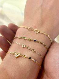 Close-up of a wrist wearing four delicate stacked bracelets: a paw-print and cutout heart chain, a multicolored bezel-gem chain, a thin script word charm, and a pavé heart with small colored stones on cream bedding.