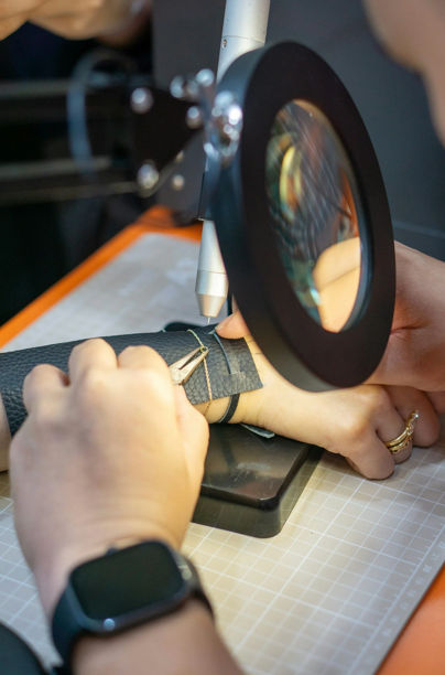 Close-up of artisan hand-stitching black leather under a magnifying lamp on a craft cutting mat