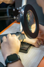 Close-up of artisan hand-stitching black leather under a magnifying lamp on a craft cutting mat