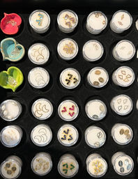 Overhead view of a black foam display tray with rows of clear round containers holding tiny gold, gemstone and crescent moon jewelry findings, plus three colorful heart-shaped dishes.
