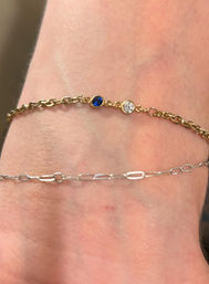 Close-up of a wrist wearing two delicate bracelets: a gold chain with small blue sapphire and clear diamond bezels paired with a thin silver paperclip-link chain.