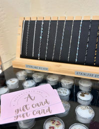 Boutique jewelry display: delicate sterling silver and stainless steel chains hanging on a wooden rack above a glass case of small earring pots, with a pink gift card in the foreground.