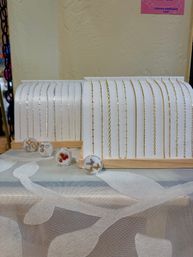 Boutique-style display of delicate gold and silver chain bracelets and necklace chains on white stands with small clear charm containers on a patterned tablecloth.