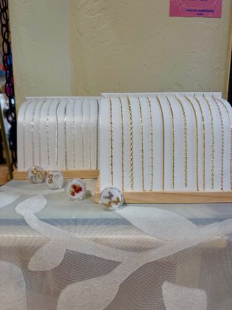 Boutique-style display of delicate gold and silver chain bracelets and necklace chains on white stands with small clear charm containers on a patterned tablecloth.