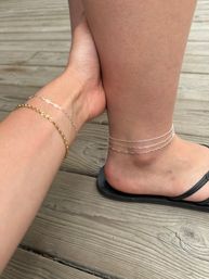 Close-up of a foot in a black flip-flop wearing layered delicate silver chain anklets, with a wrist nearby wearing thin gold and silver bracelets, photographed on a weathered wooden deck.