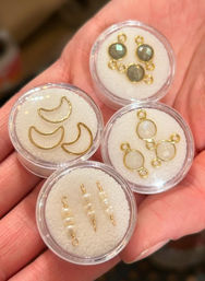 Hand holding five clear round cases of jewelry-making components: gold crescent charms, iridescent round gemstone bezels, gold-loop moonstone-style connectors, and small wired freshwater pearl bar connectors — DIY earring and pendant supplies.