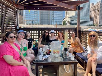 Eight friends in swimsuits smiling in a wooden rooftop cabana by an urban waterfront with skyscrapers, table with fruit-infused water and drinks — summer city gathering
