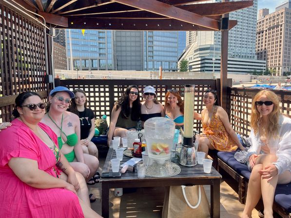 Eight friends in swimsuits smiling in a wooden rooftop cabana by an urban waterfront with skyscrapers, table with fruit-infused water and drinks — summer city gathering
