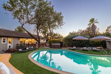 Dusk view of a suburban backyard with a curved turquoise swimming pool, lounge chairs and scalloped umbrellas on green turf, a hot tub, string lights and large trees beside a single-story house.