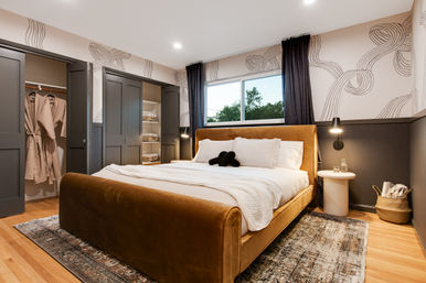 Cozy modern master bedroom with caramel velvet upholstered bed, white linens and black accent pillows, patterned wallpaper, dark gray built-in closets with hanging robes, round nightstand and lamp, woven basket, hardwood floors and area rug with a window showing treetops.