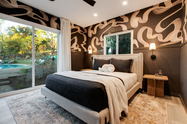 Sunlit modern bedroom with bold abstract wallpaper, upholstered bed in black-and-white linens, bedside sconces, and a sliding glass door opening to a backyard pool and trees.