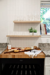 Modern kitchen vignette: wooden butcher block island with a charcuterie board, wine bottle and glasses, floating wood shelves holding clear glassware and small potted herbs, cookbooks by a sunlit window and granite countertops.