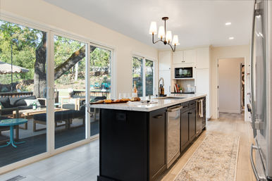 Sunlit open-concept kitchen with a large black island and white marble countertop, stainless steel appliances, pendant chandelier, and sliding glass doors leading to a shaded backyard deck with outdoor seating and oak trees.