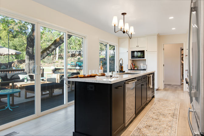 Sunlit open-concept kitchen with a large black island and white marble countertop, stainless steel appliances, pendant chandelier, and sliding glass doors leading to a shaded backyard deck with outdoor seating and oak trees.