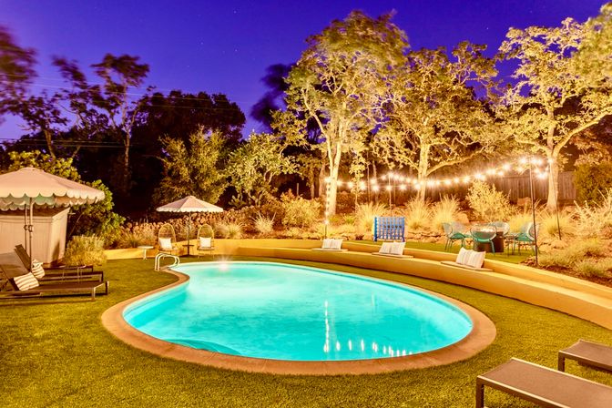 Evening backyard with an illuminated kidney-shaped turquoise pool, artificial-turf lawn, lounge chairs and umbrellas, string lights draped through trees, and cozy patio seating in a landscaped garden.