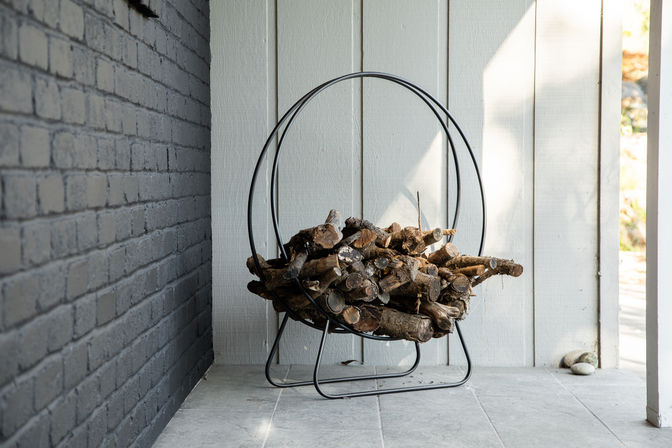 Round black metal firewood rack holding chopped logs on a tiled porch beside gray brick and white wood siding, sunlight casting soft shadows