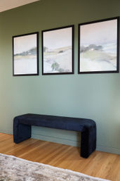 Green accent wall with three black‑framed abstract landscape prints above a navy upholstered bench on hardwood floor with a neutral rug, modern entryway vignette.