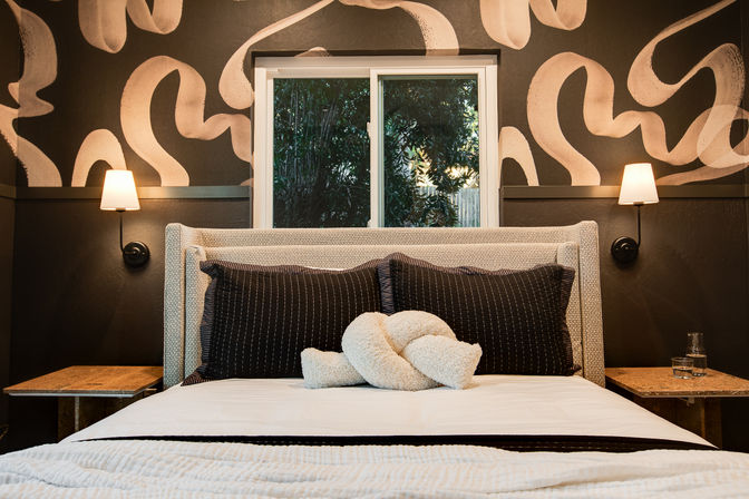 Cozy modern bedroom with upholstered headboard, black pinstriped pillows and a cream knot cushion on white bedding, flanked by wall sconces and a bold abstract accent wall with a centered window showing garden foliage.
