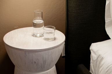 Round whitewashed bedside table topped with a clear water carafe and tumbler, beside a dark upholstered headboard and white bedding against a beige wall.