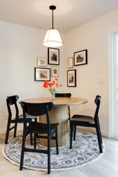 Bright corner dining nook with round wood table, four black chairs, vase of orange flowers on a patterned round rug under a pendant light and framed gallery wall