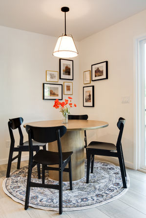 Bright corner dining nook with round wood table, four black chairs, vase of orange flowers on a patterned round rug under a pendant light and framed gallery wall