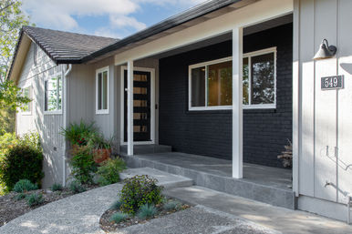 Modern suburban front porch with a black glass-panel door, dark brick accent wall, gray siding, tiled steps and a landscaped curved walkway.