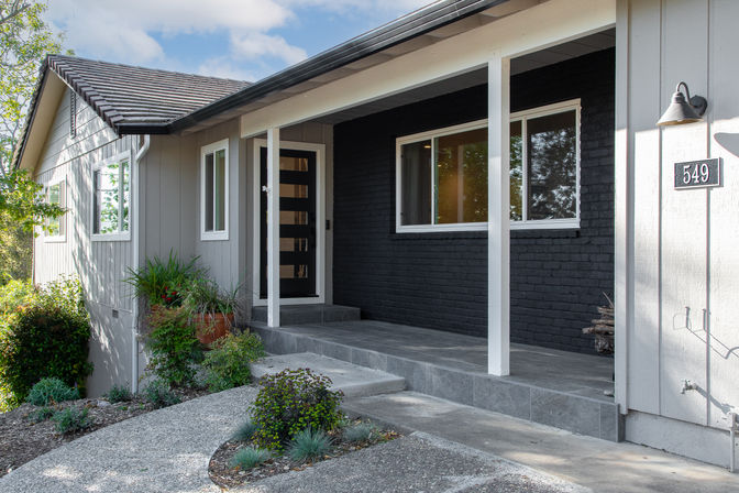 Modern suburban front porch with a black glass-panel door, dark brick accent wall, gray siding, tiled steps and a landscaped curved walkway.