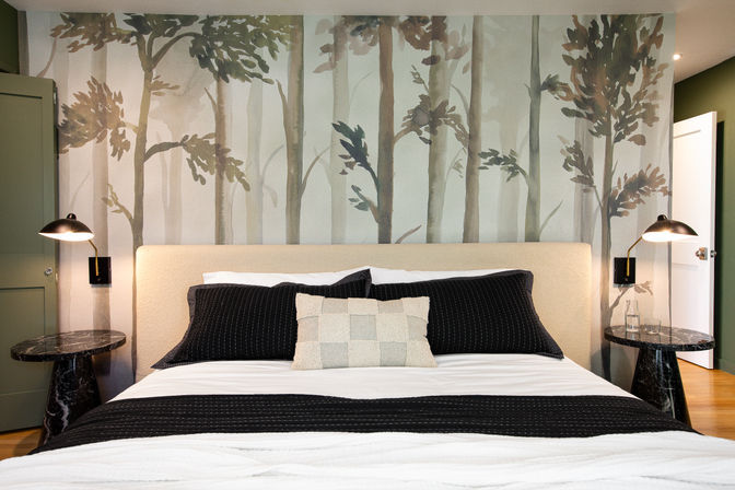 Contemporary cozy bedroom interior with upholstered headboard, black pillows and throw, forest mural wallpaper, marble nightstands and brass wall lamps.