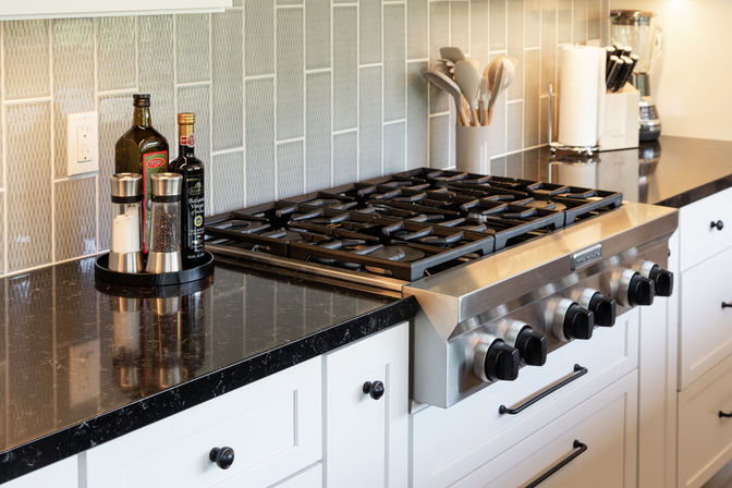 Sleek modern kitchen gas cooktop with stainless-steel range and black quartz countertops, light textured tile backsplash, white shaker cabinets, and olive oil, vinegar and salt-and-pepper set on the counter.