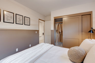 Cozy modern bedroom interior with white bedding and round textured pillow, two-tone neutral walls with framed botanical prints, and a beige sliding-door closet holding hanging robes.