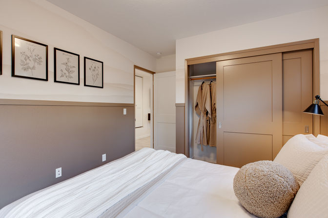 Cozy modern bedroom interior with white bedding and round textured pillow, two-tone neutral walls with framed botanical prints, and a beige sliding-door closet holding hanging robes.