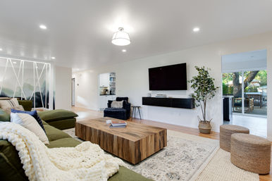 Bright modern open-concept living room with a green sectional and chunky knit throw, low wooden coffee table, wall-mounted flat-screen TV, navy armchair, woven poufs, potted indoor plant, and sliding doors to a tree-filled patio.