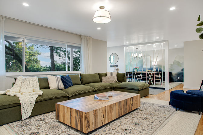 Bright modern open-concept living room with olive-green sectional, chunky knit throw, wood slab coffee table on patterned rug, large picture window framing leafy trees, and a dining area visible through a decorative white room divider.