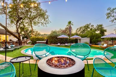 Dusk backyard pool patio with glowing round fire pit, turquoise mesh chairs, string lights, lounge umbrellas and palm-framed trees.