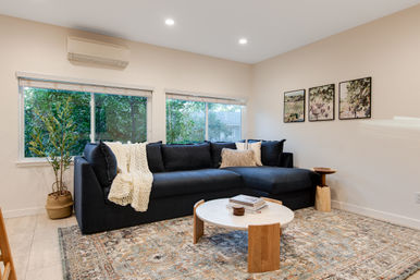 Cozy modern living room with a navy L-shaped sectional draped in a chunky cream throw, round marble coffee table on a patterned area rug, potted plant by wide windows with garden views, and three botanical wall prints.